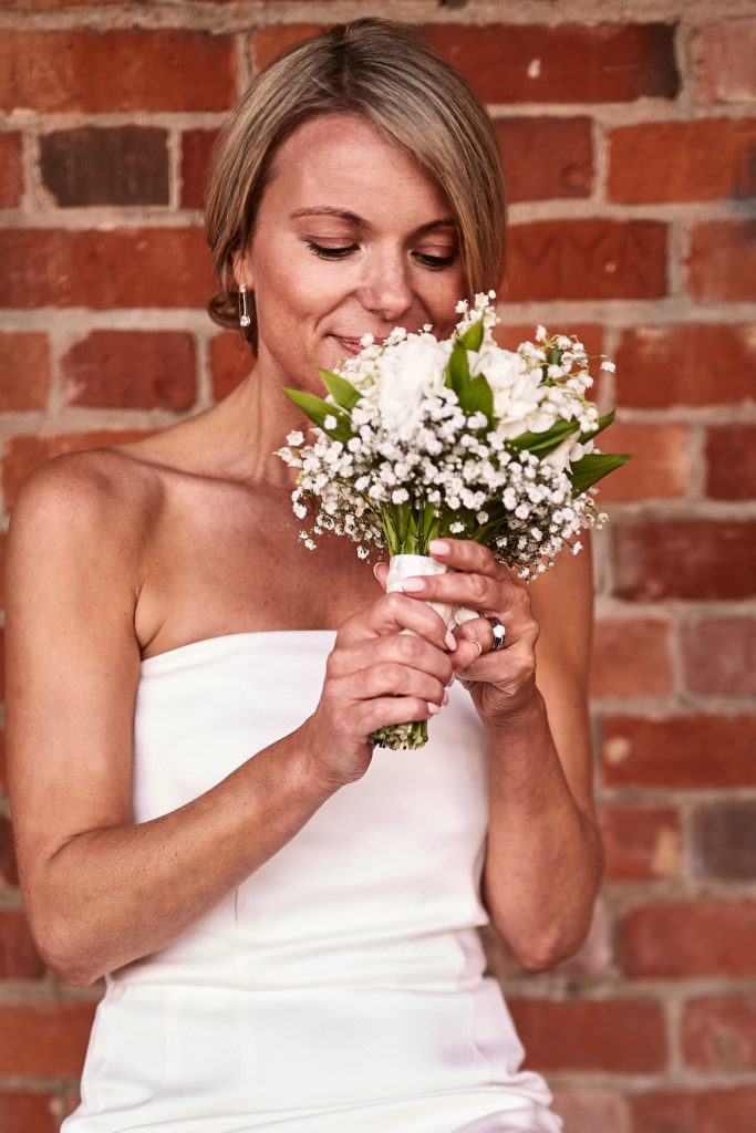 Bride smelling her bouquet of white flowers against a red-brick wall