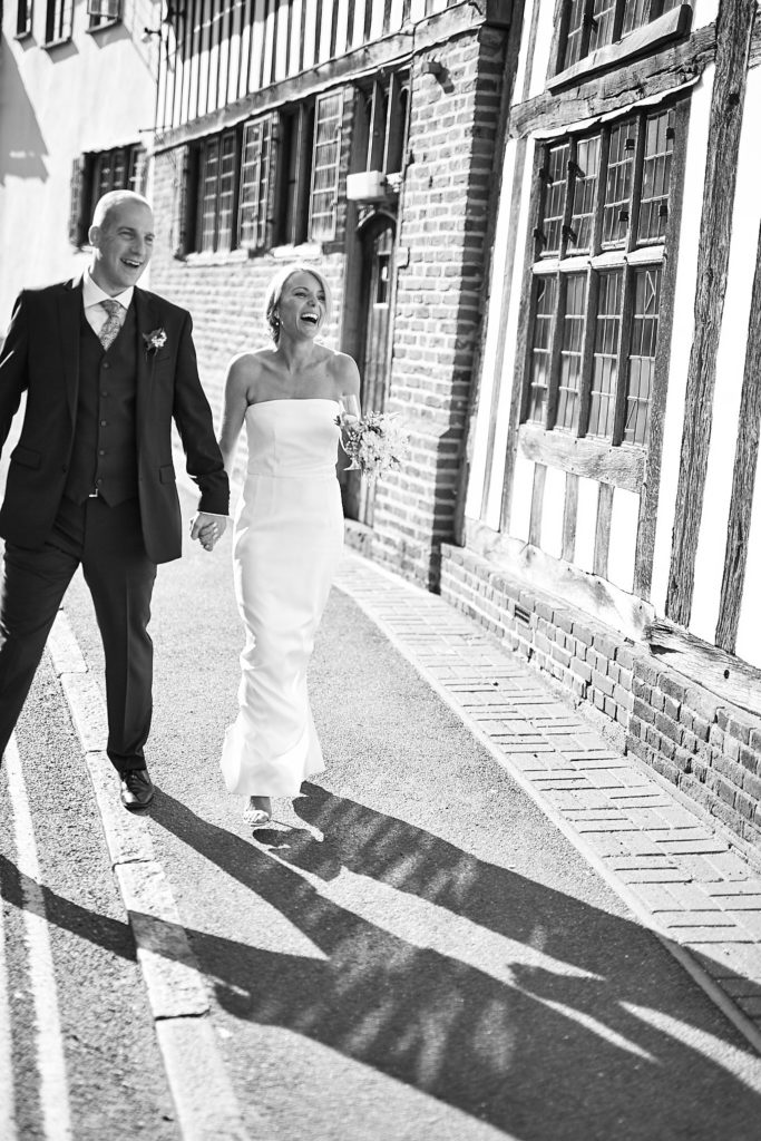 Black and white photo of the bride and groom walking hand in hand past a timber-framed building in Lavenham