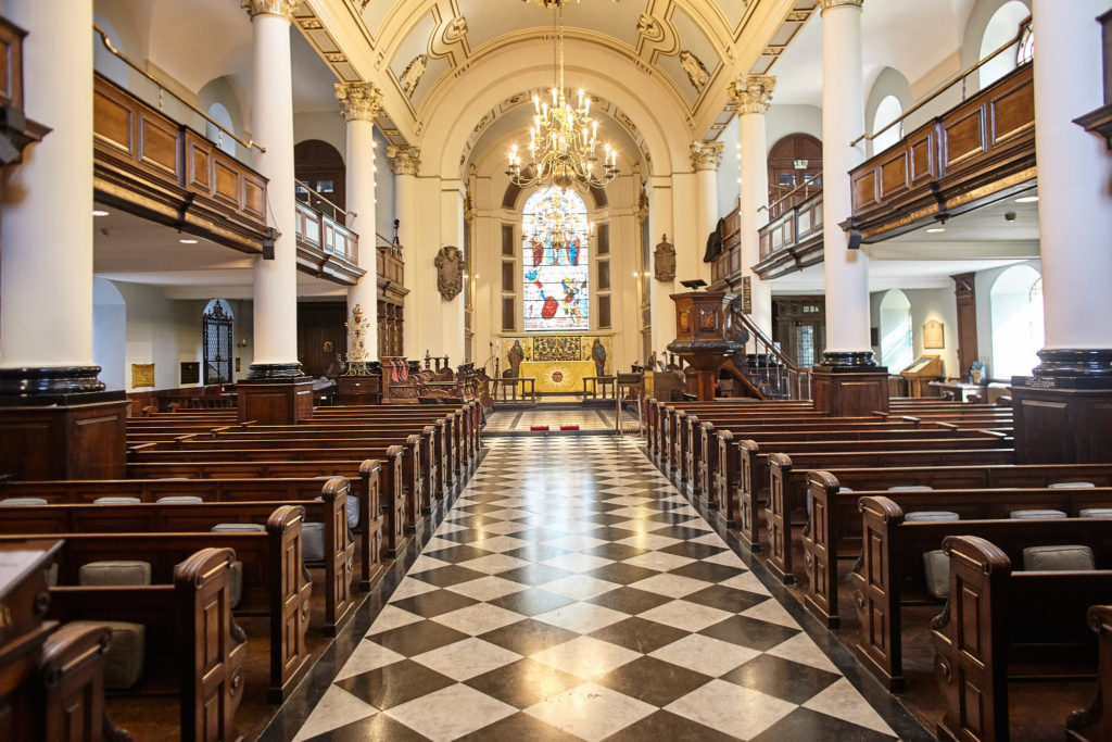 Interior of a London church with a chequered floor and wooden pews, ready for the wedding