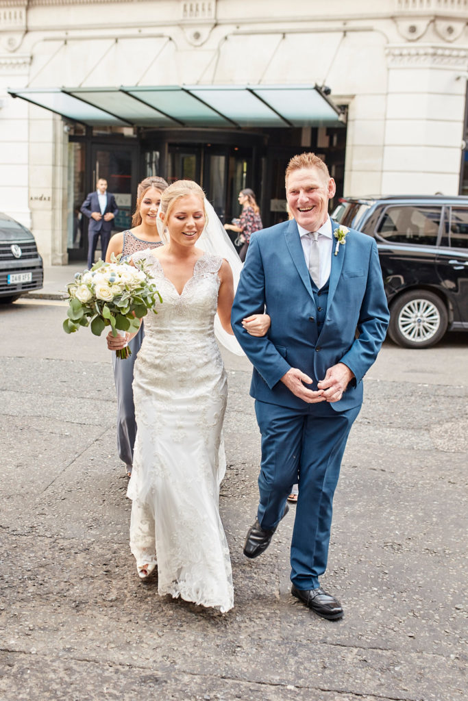 Bride walking to the church arm in arm with a man in a blue suit, outside the Andaz in London