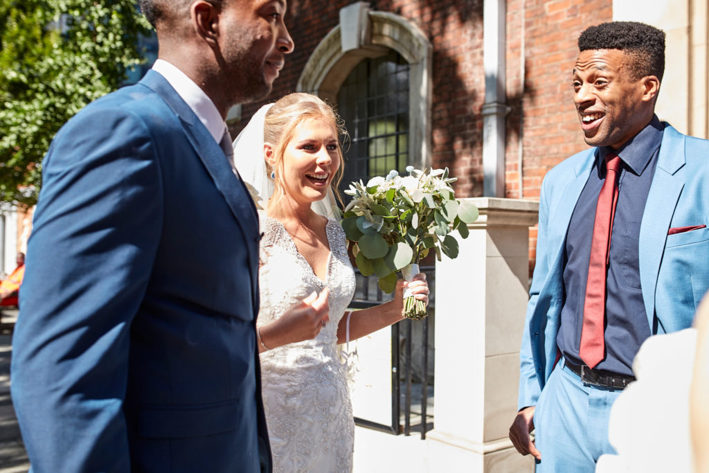 Bride and groom laughing with a guest in a light blue suit outside the church