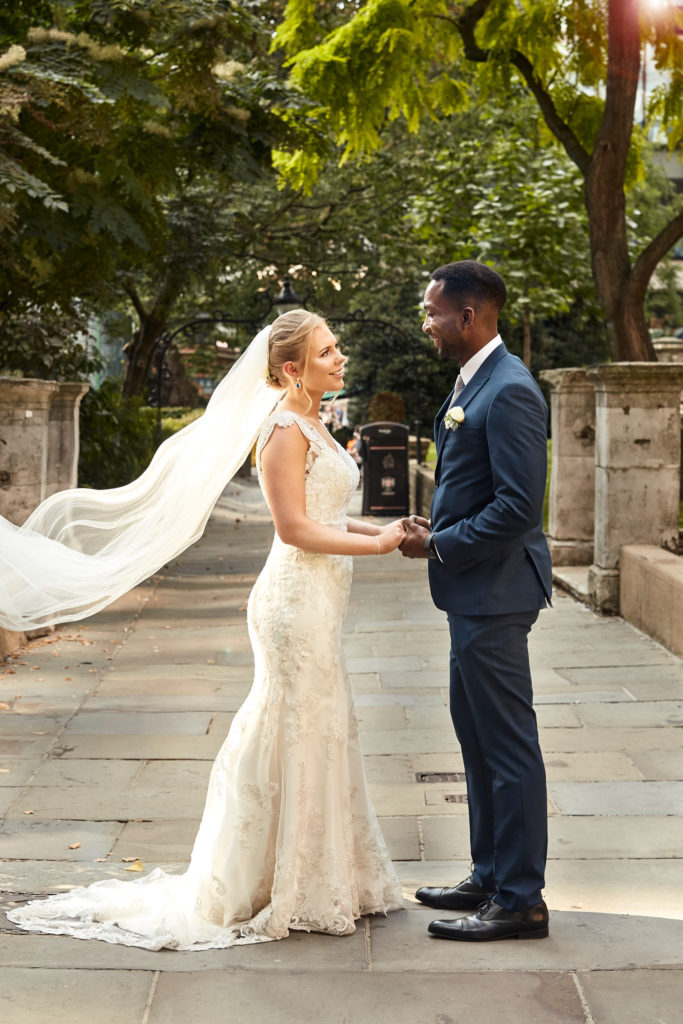 Bride and groom holding hands on a paved path as her veil blows out behind her