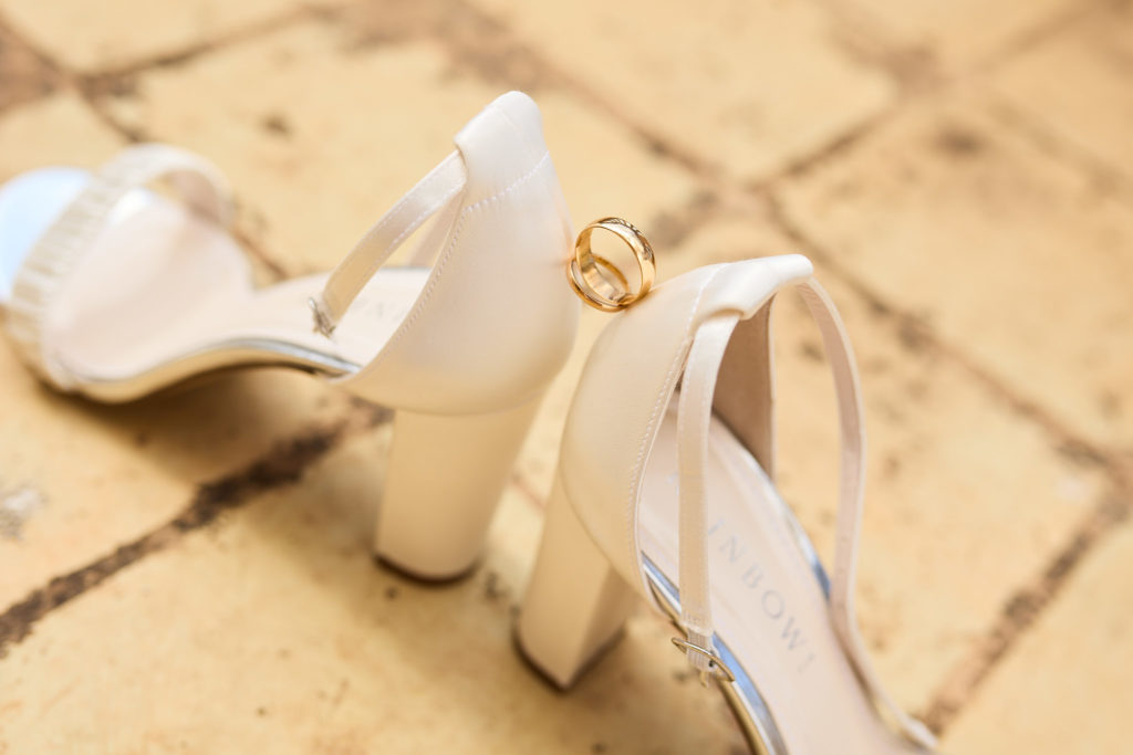 White block-heel bridal shoes with a wedding ring on a tiled floor