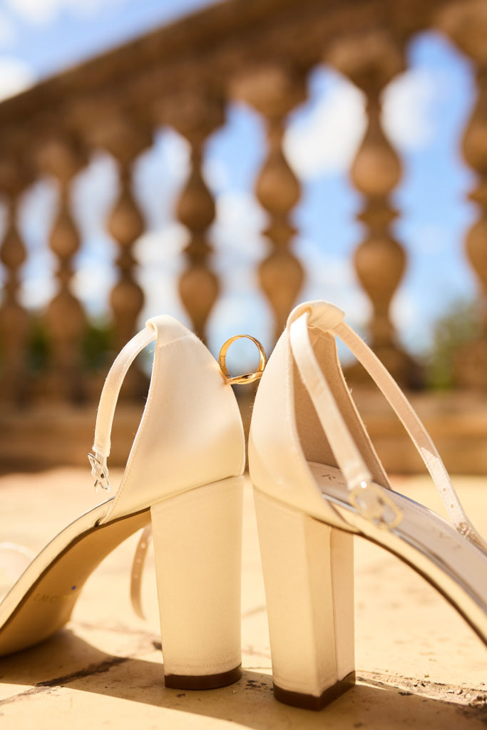 White bridal shoes with a ring balanced between them in front of a stone balustrade