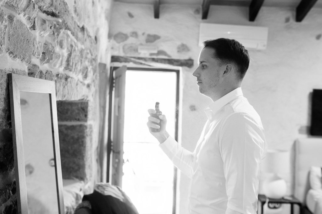 Black and white photo of the groom getting ready in a white shirt beside a window