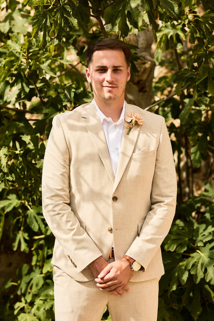 Groomsman in a beige suit with a white buttonhole in front of fig leaves