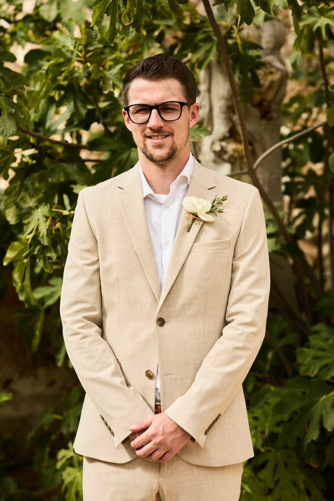 Groomsman in glasses and a beige suit smiling in front of fig leaves