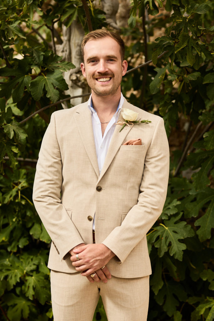 Groomsman with a moustache in a beige suit smiling in front of fig leaves