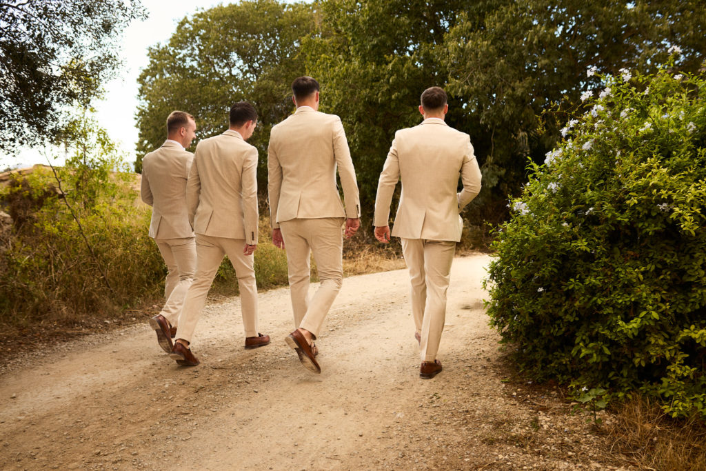 Groom and three groomsmen in beige suits walking along a sandy track at Son Boronat, seen from behind