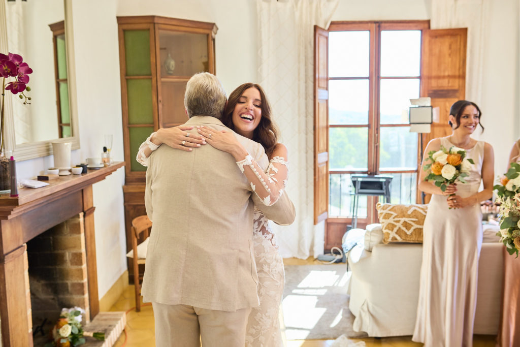 Man in a beige suit hugging the bride as a bridesmaid looks on