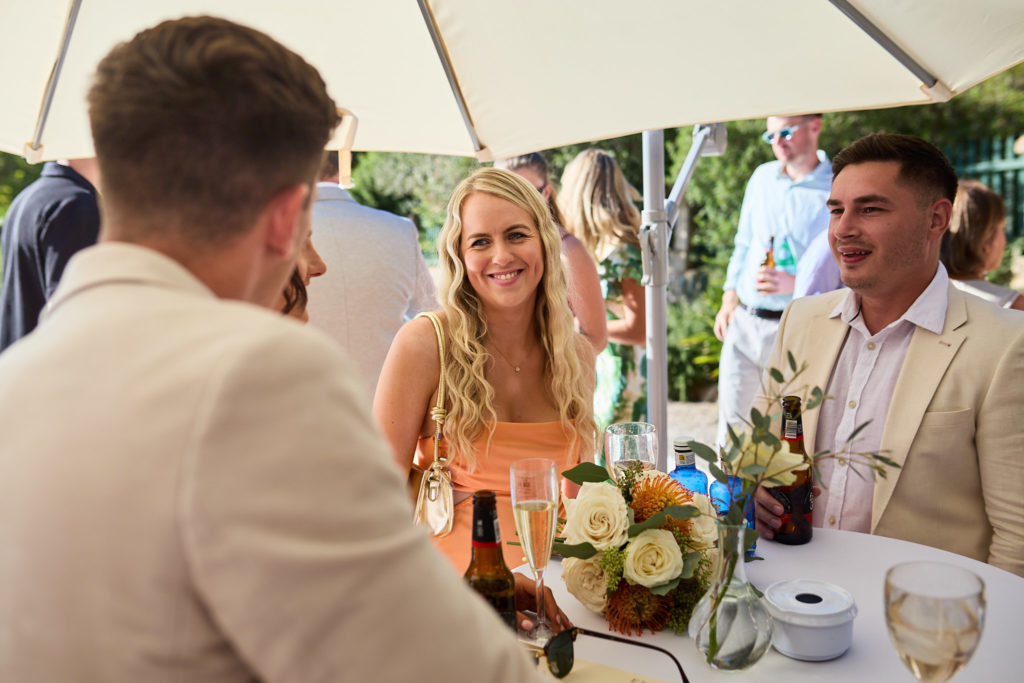 Wedding guests chatting at a table in the shade beside the pool