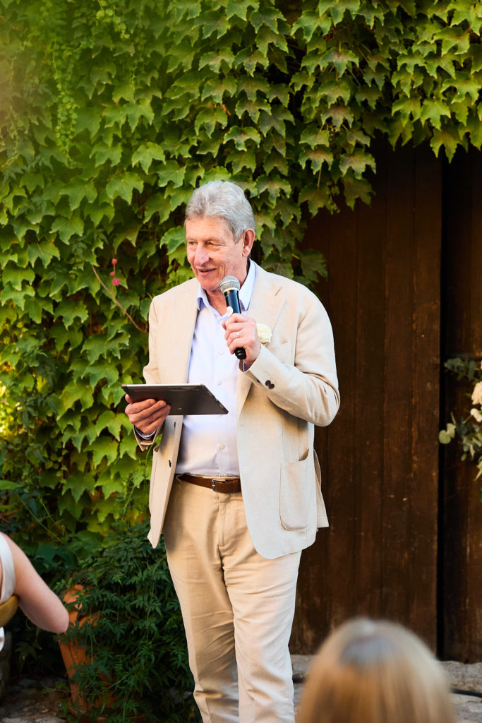 Older man in a beige suit giving a speech with a microphone