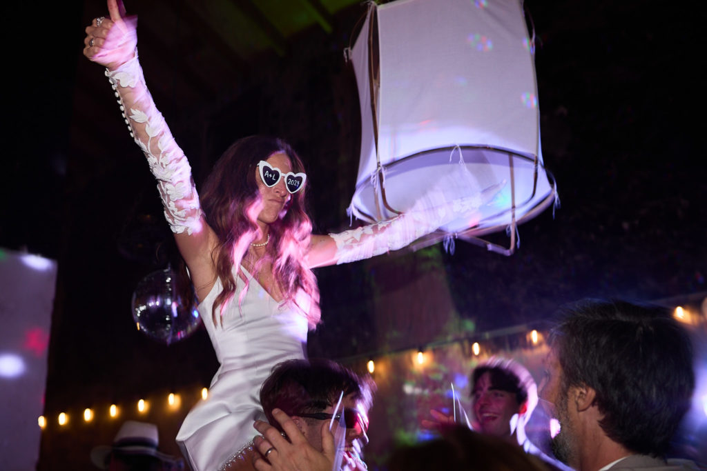 Bride in sunglasses on someone's shoulders waving her arm above the dance floor