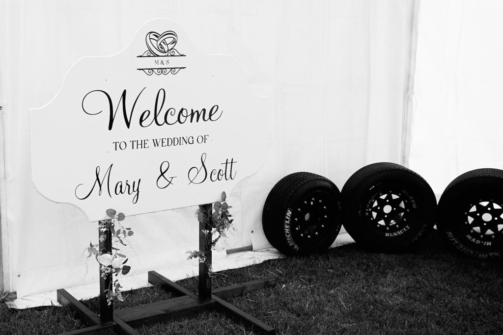 Black and white welcome sign for Mary and Scott's wedding beside a stack of car tyres