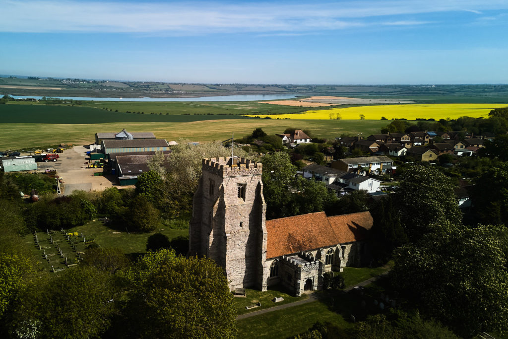 Aerial drone photo of St Nicholas Church in Canewdon, near Rochford, Essex, with yellow fields and the River Crouch beyond