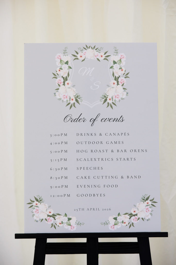 Order of events sign with a floral wreath on an easel in the marquee