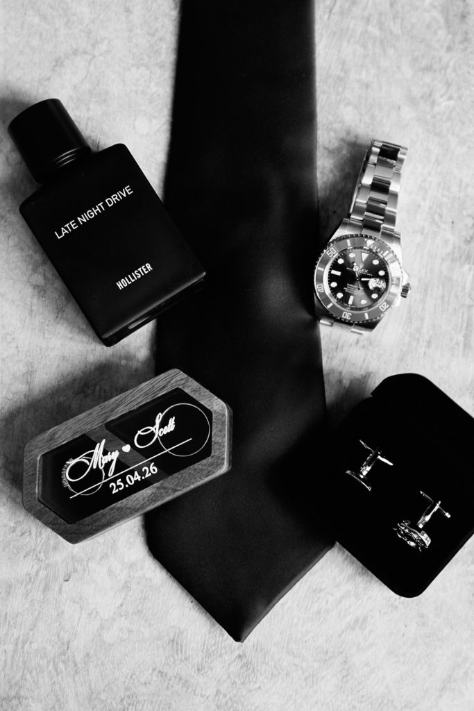 Black and white flat lay of the groom's tie, watch, aftershave and cufflinks