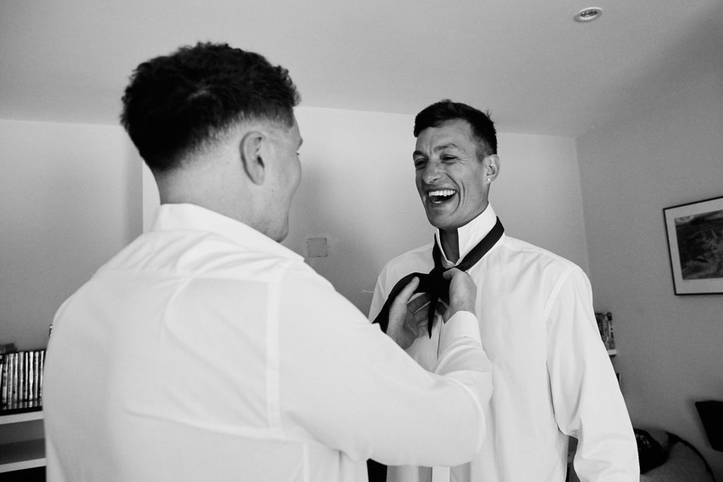 Black and white photo of a groomsman straightening the groom's tie as they laugh