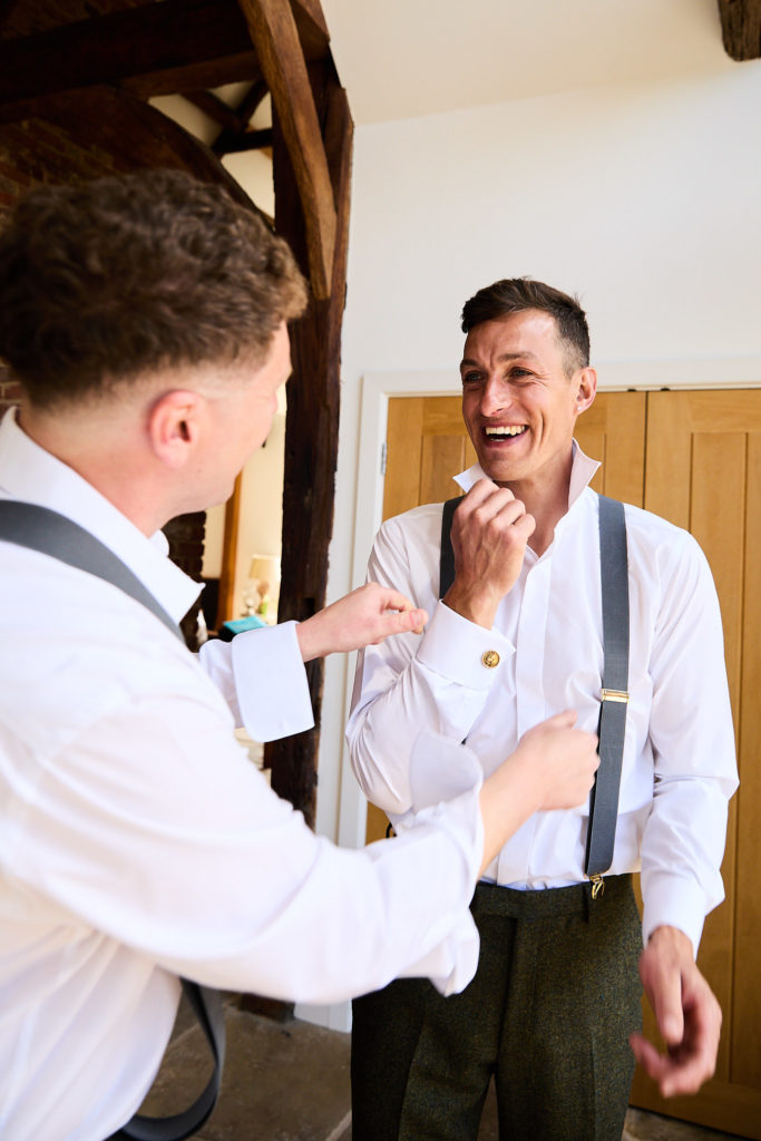 Groom and groomsman laughing as they get dressed in shirts and braces