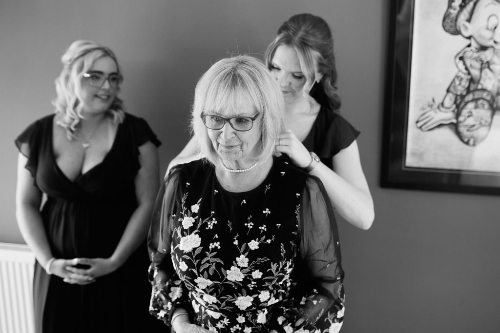 Black and white photo of a woman having her necklace fastened before the wedding