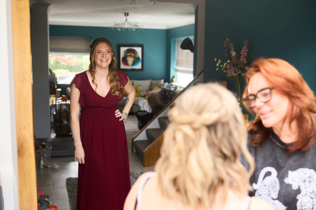 Bridesmaid in a burgundy dress smiling across the room at the bride during bridal prep