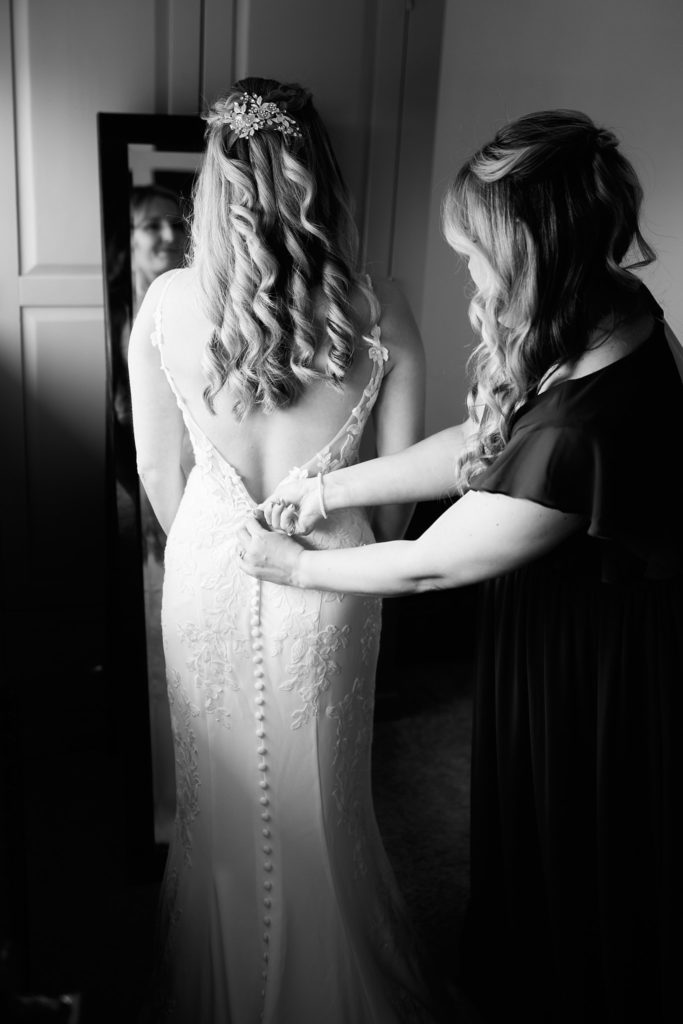 Black and white photo of a bridesmaid buttoning the back of the bride's dress