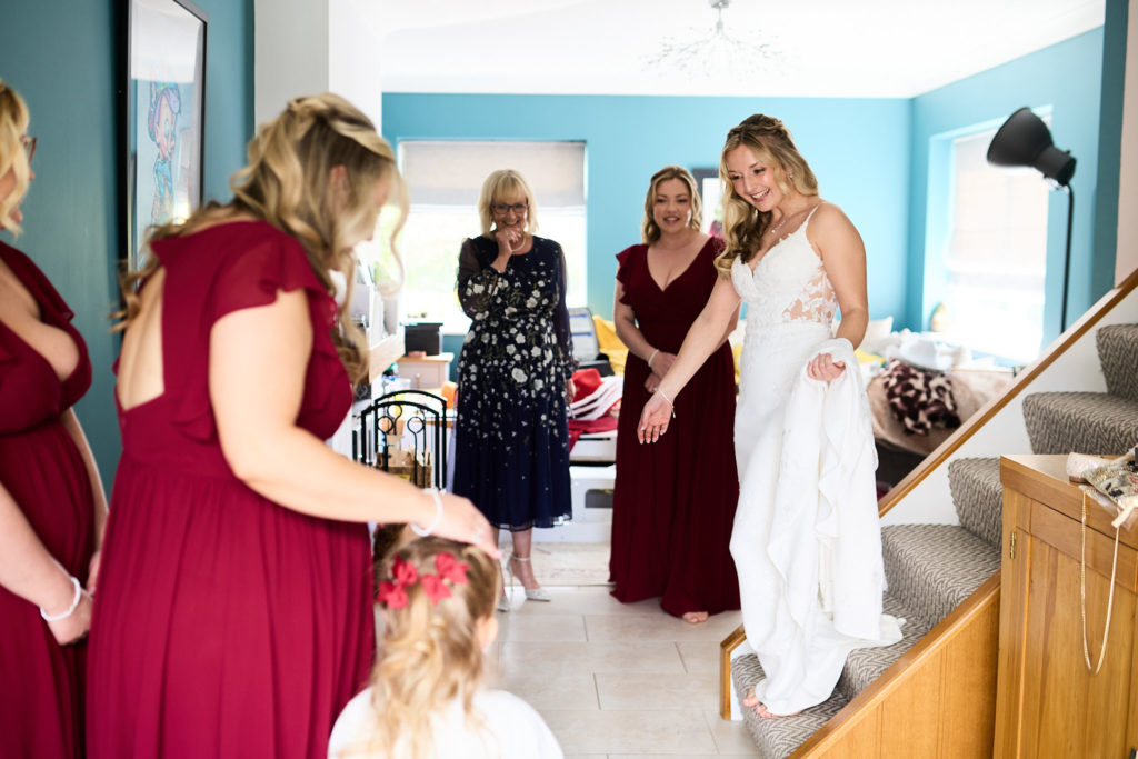 Bride in her wedding dress at the foot of the stairs as bridesmaids and family look on