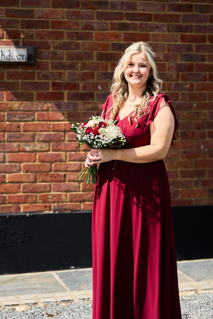 Smiling bridesmaid in a burgundy dress with a red and white bouquet by a brick wall