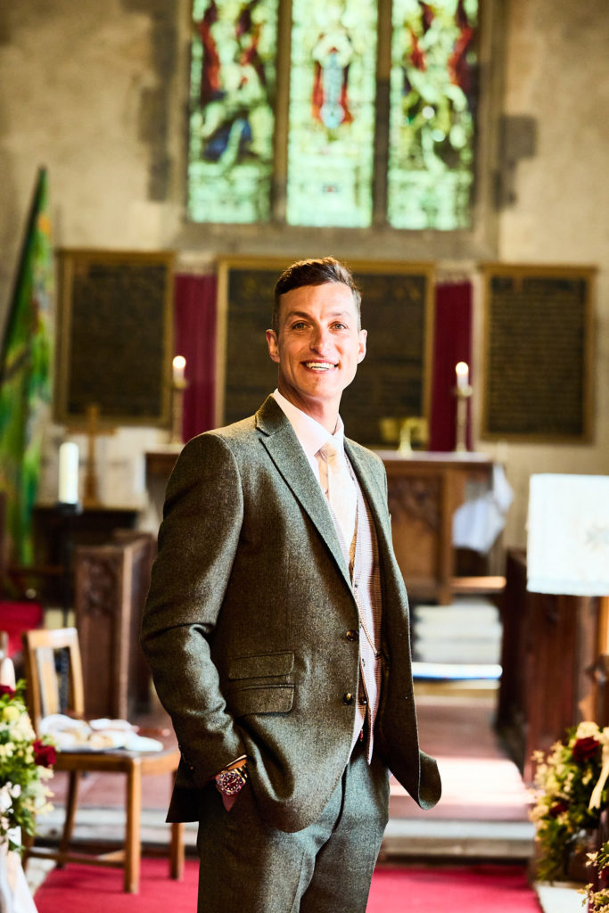 Groom waiting in St Nicholas Church, Canewdon, in front of a stained glass window