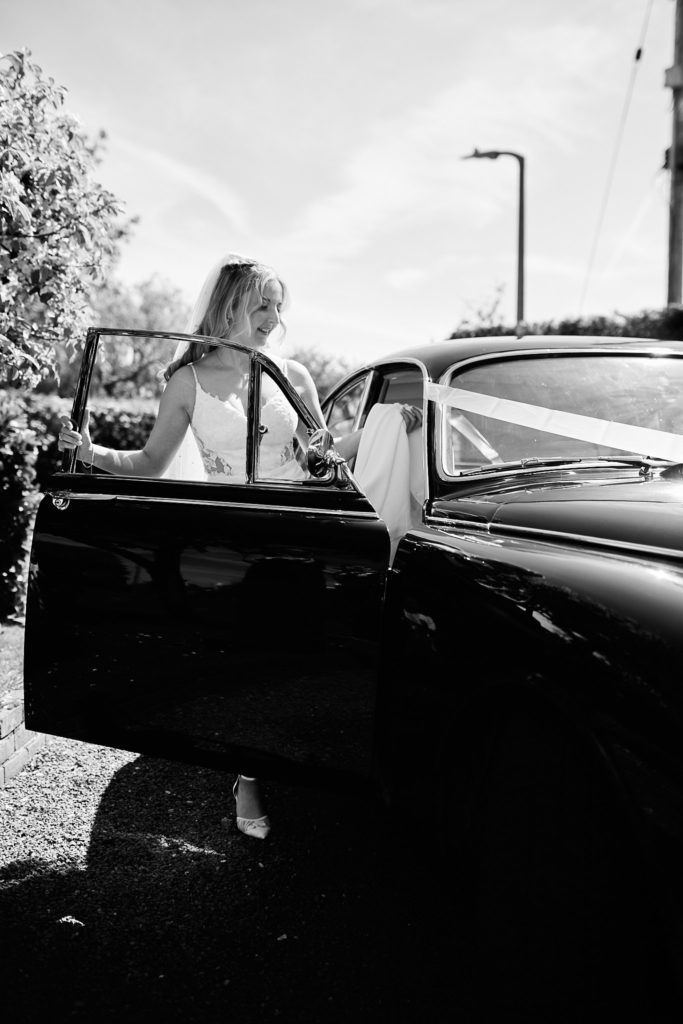 Black and white photo of the bride getting into a classic wedding car tied with ribbon