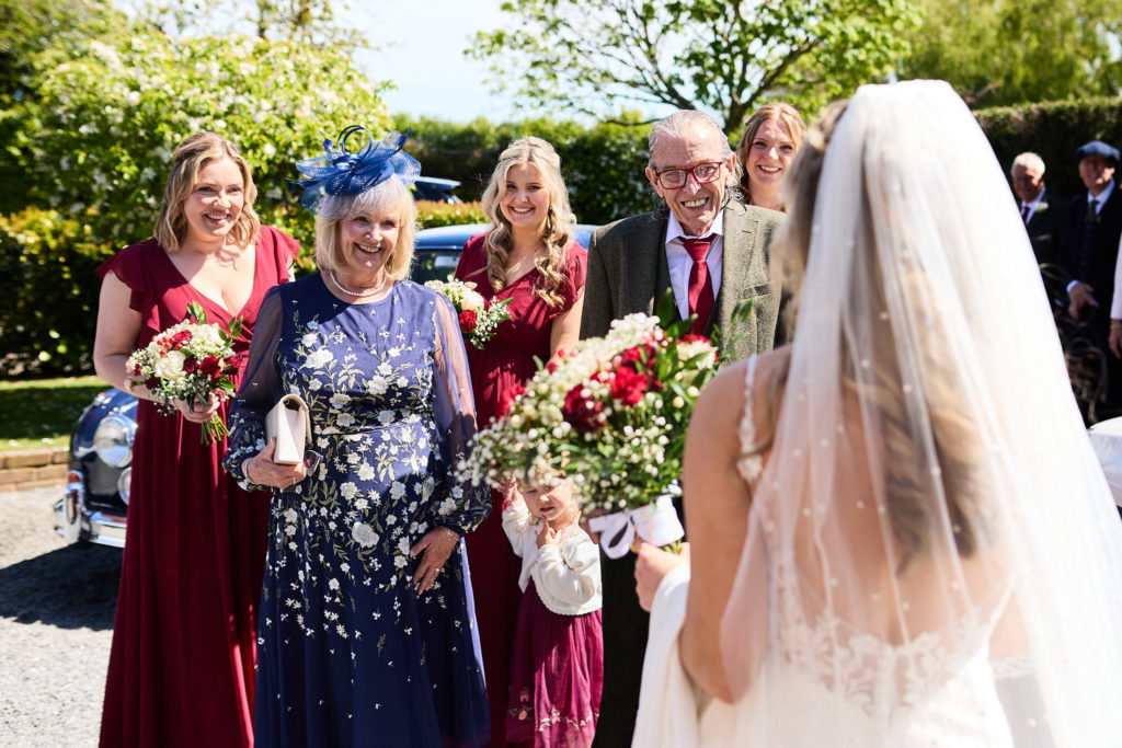 Bride in her veil, seen from behind, greeting smiling bridesmaids and family outside the church