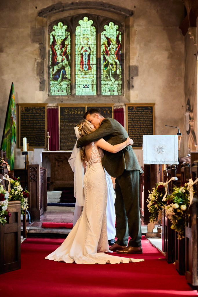 Bride and groom hugging at the altar beneath a stained glass window