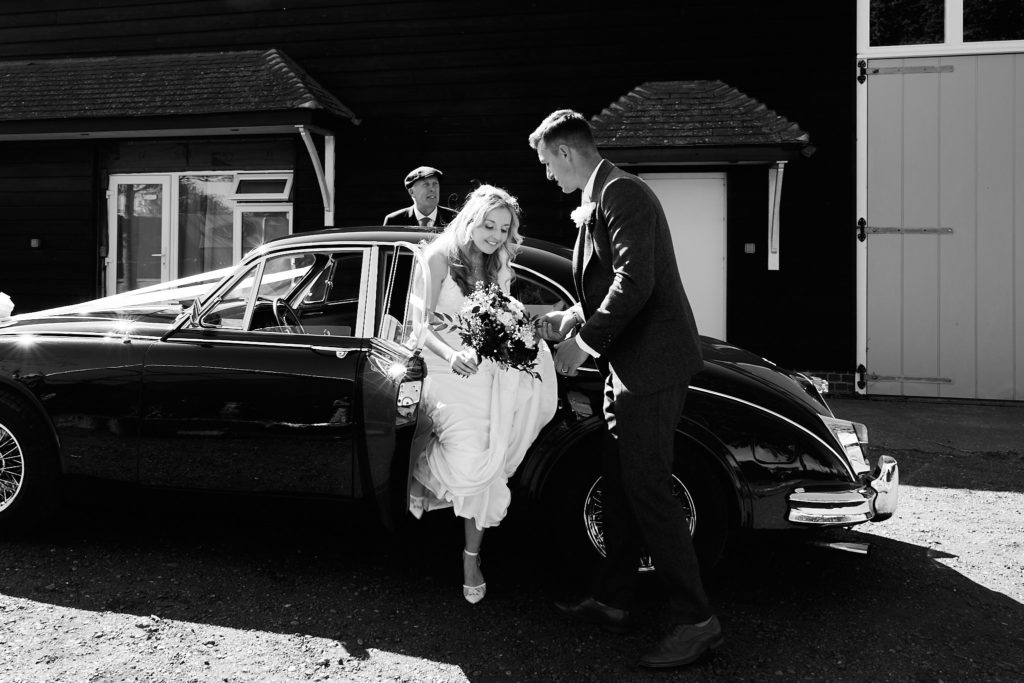 Black and white photo of the groom helping the bride out of the classic wedding car