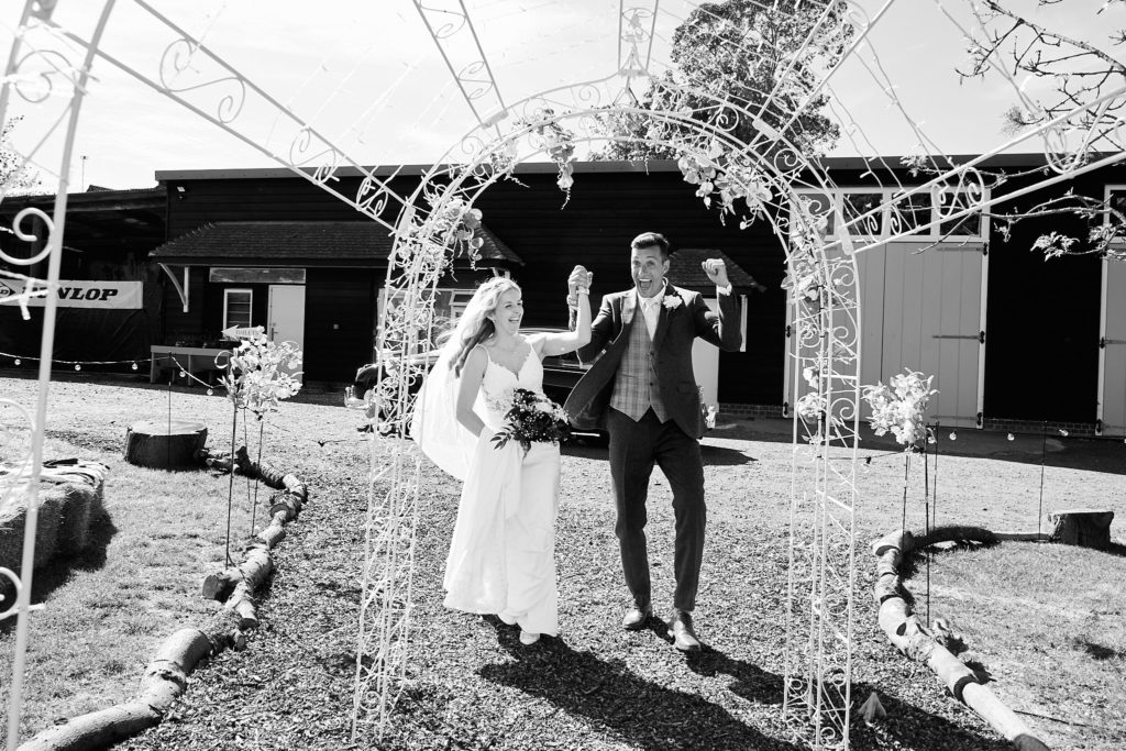 Black and white photo of the bride and groom waving as they walk under a floral arch