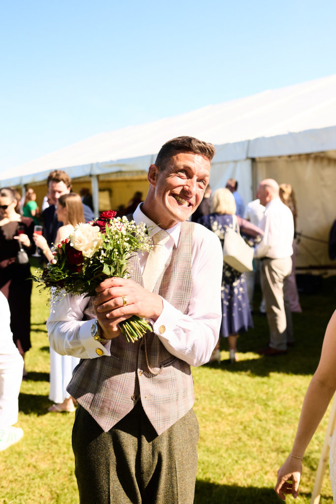 Man in a waistcoat laughing as he holds the bouquet outside the marquee