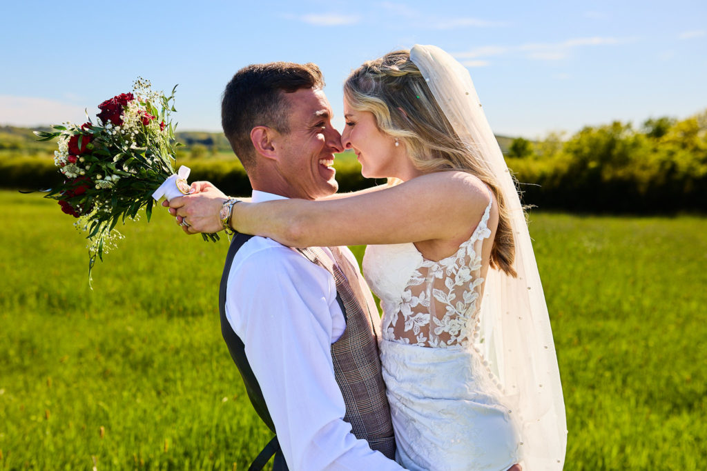 Bride laughing with her arms round the groom's neck in a sunny meadow near the River Crouch