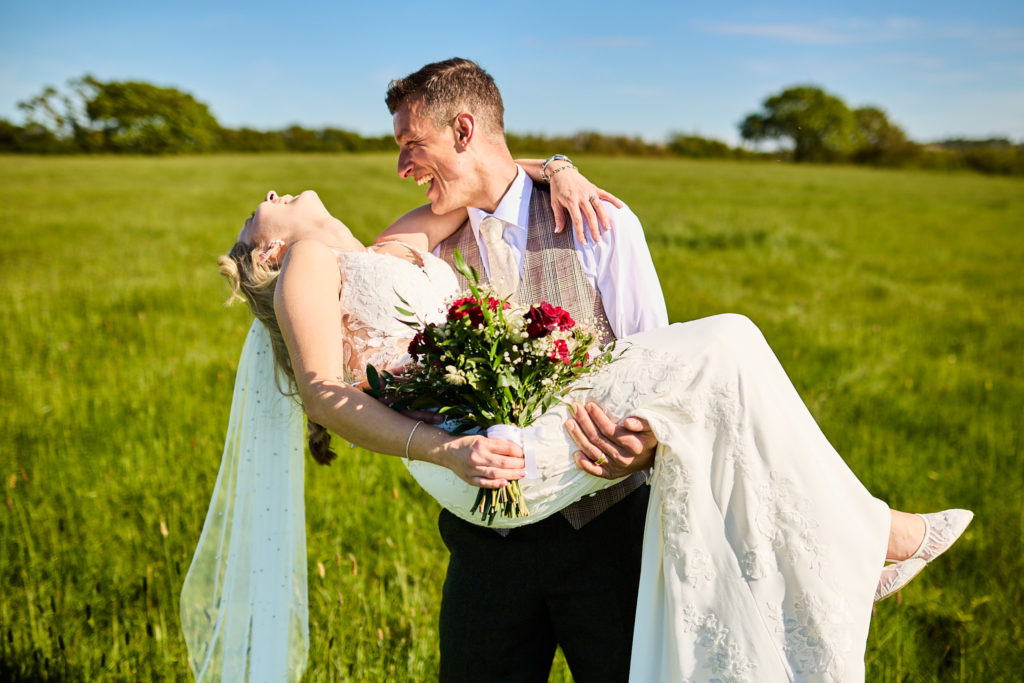 Groom carrying the bride through a field, her veil trailing