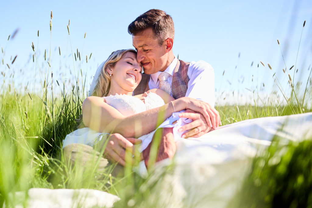Bride and groom cuddling in long grass under a blue sky
