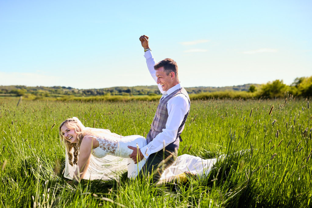 Groom punching the air beside the bride sitting in long grass