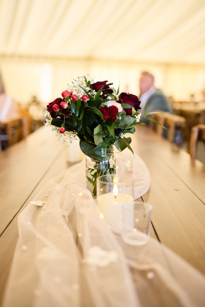 Jar of red roses on a long wooden table in the wedding marquee