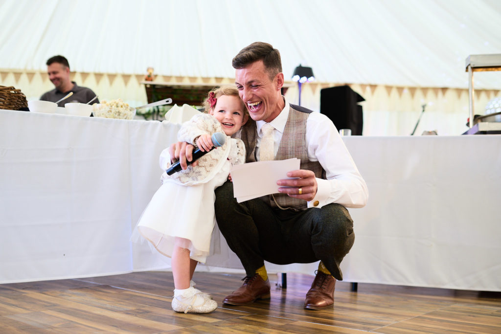 Groom crouching beside a flower girl holding the microphone
