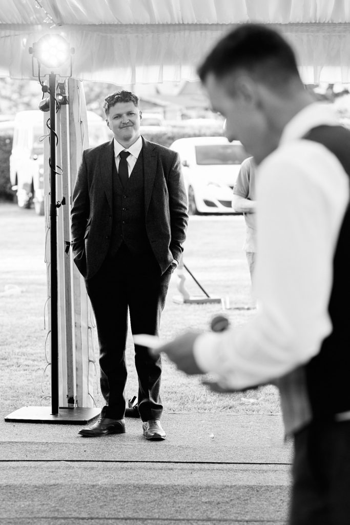 Black and white photo of the groom giving his speech as a man stands listening