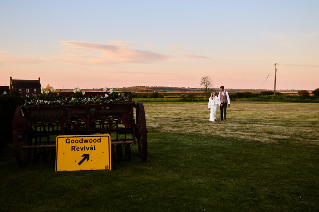 Bride and groom standing in a field at dusk beyond a yellow arrow sign
