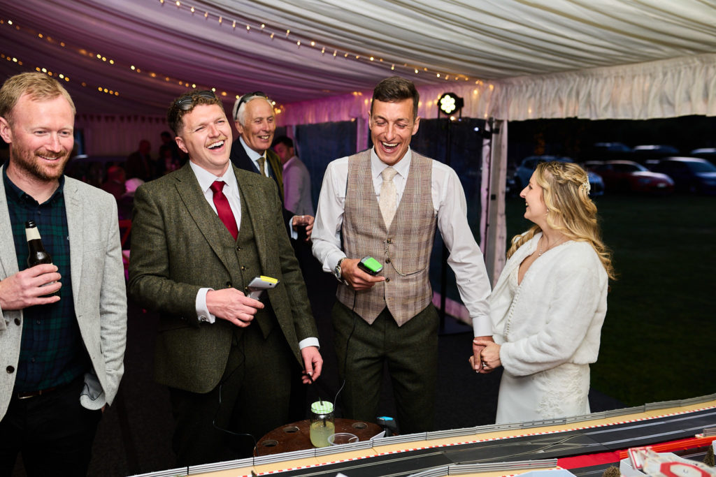 Bride, groom and guests laughing around the Scalextric track in the evening