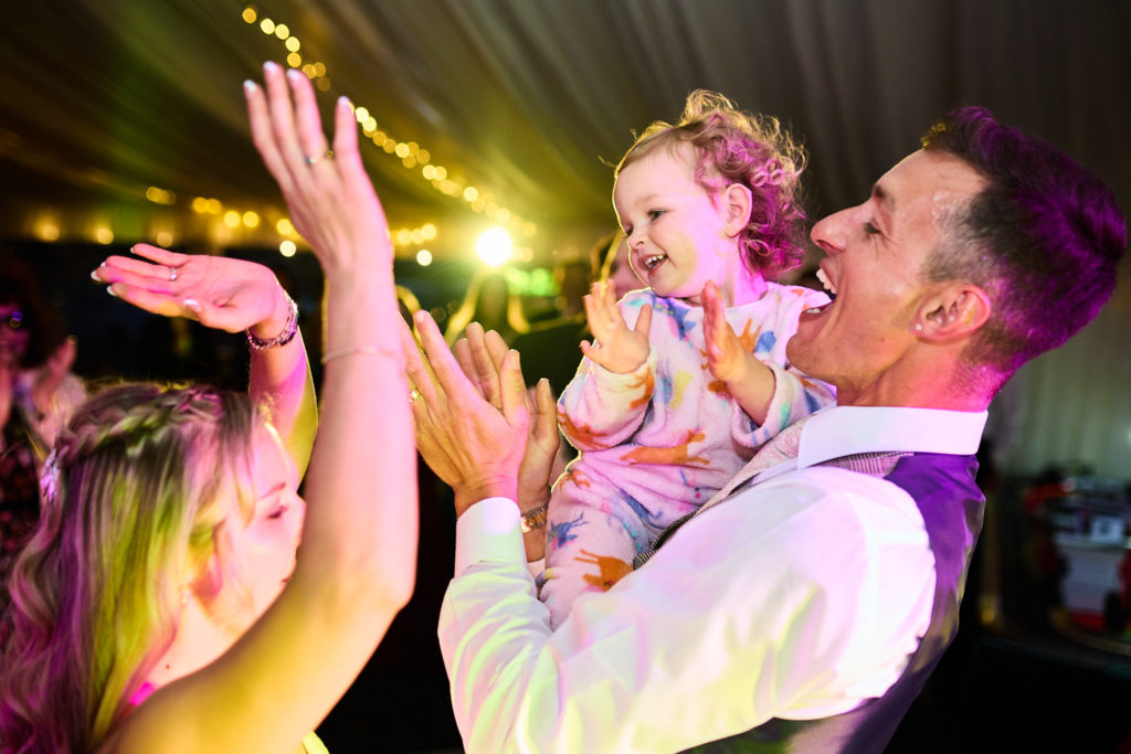 Groom holding a toddler as guests clap under festoon lights