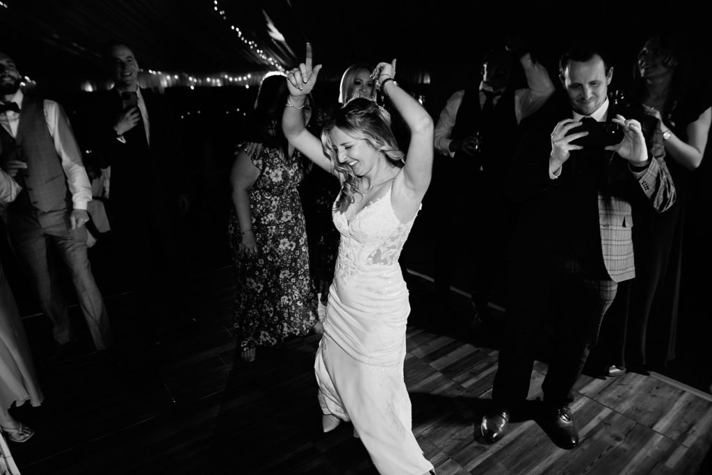 Black and white photo of the bride dancing with her arms raised as a guest films