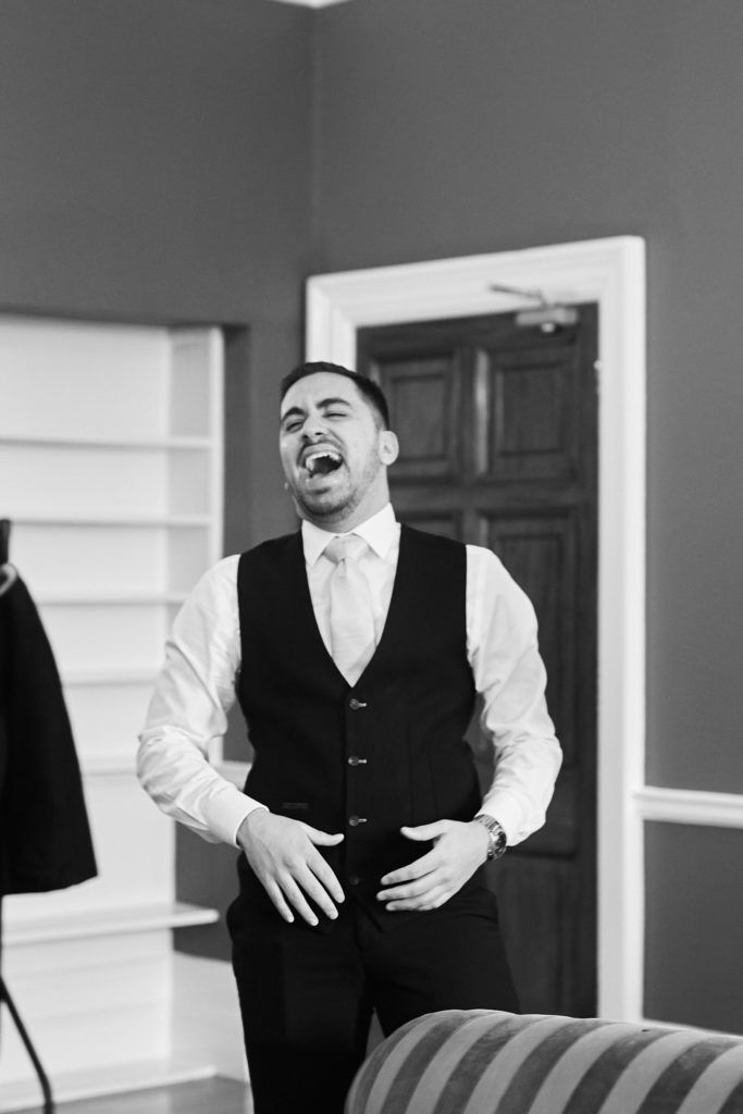 Black and white photo of a groomsman laughing in a waistcoat as he gets ready
