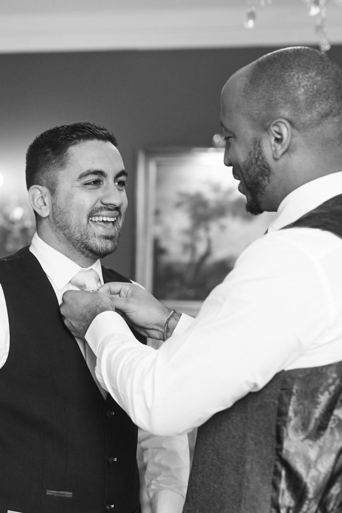 Black and white photo of the groom fixing a groomsman's tie as they laugh