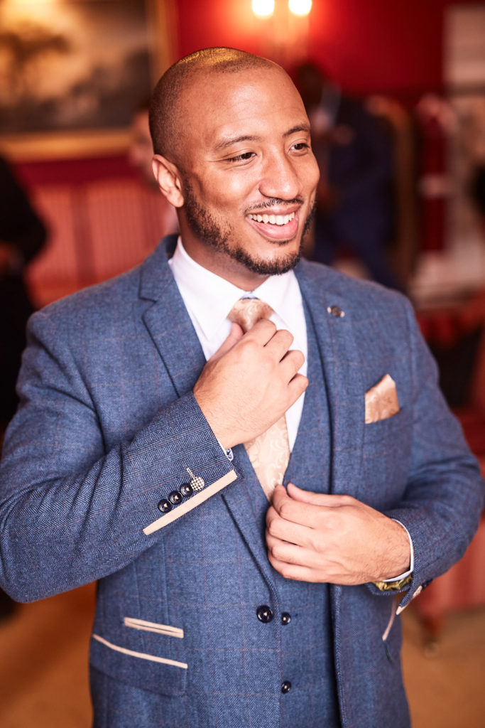 Groom smiling as he straightens his tie in a blue tweed three-piece suit