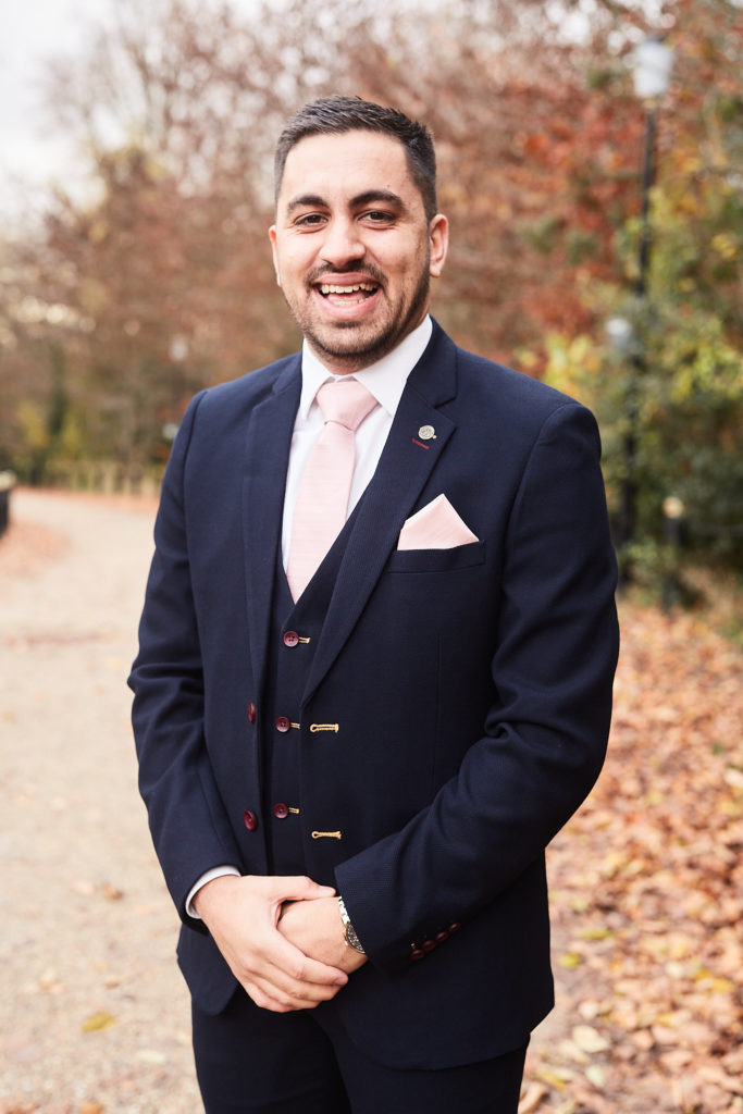 Groomsman smiling in a navy three-piece suit with a blush tie on an autumn drive
